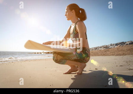 woman surfer kneeling on the beach with her surfboard watching the waves, lens flare