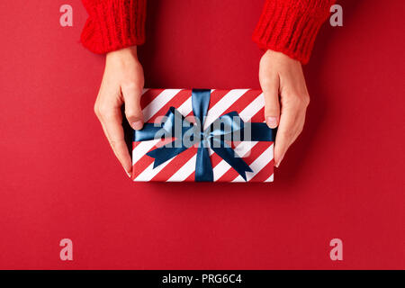 Female's hands in red pullover holding striped gift box with blue ribbon on red background. Christmas, New Year, Valentine's day and birthday concept. Stock Photo