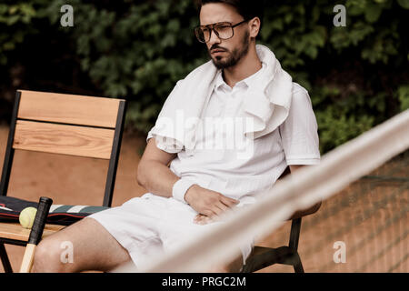 serious handsome tennis player resting on chair at tennis court Stock Photo