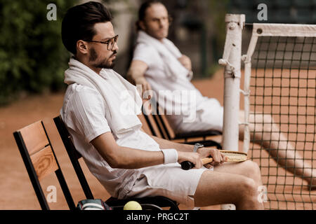 side view of retro styled tennis players resting on chairs with towels and rackets at tennis court Stock Photo