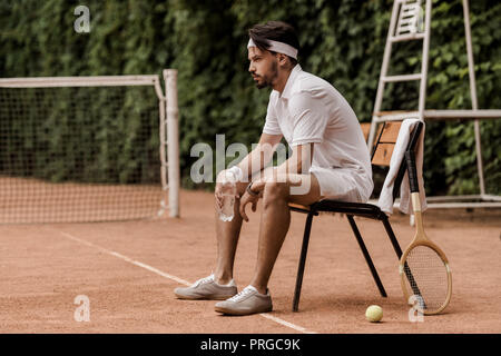 side view of handsome retro styled tennis player sitting on chair with bottle of water at tennis court Stock Photo