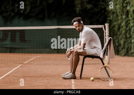 handsome retro styled tennis player sitting on chair with bottle of water at tennis court Stock Photo