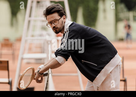 side view of handsome retro styled tennis player leaning on tennis net at court Stock Photo