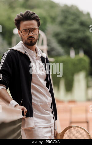 handsome retro styled tennis player leaning on tennis net at court and looking at camera Stock Photo