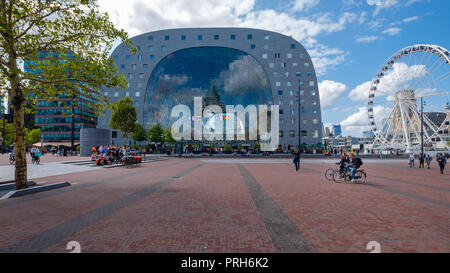 Look through with nice reflections in the glass of the market hall in Rotterdam. A Ferris wheel next to the building and many people and cyclists on t
