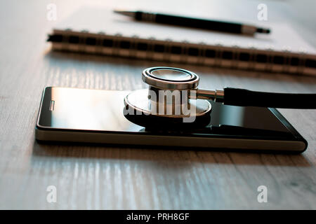 Stethoscope On A Black Smartphone Next To A Notepad And Biro - Fixing A Broken Mobile Phone Concept Stock Photo