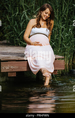 portrait of beautiful and cheerful pregnant woman sitting at the lake with her feet in the water Stock Photo