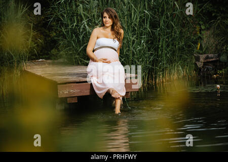 portrait of beautiful and cheerful pregnant woman sitting at the lake with her feet in the water Stock Photo