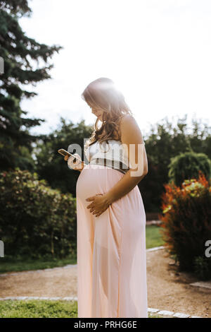 portrait of beautiful pregnant woman is holding her belly and looking at an app at her smartphone Stock Photo