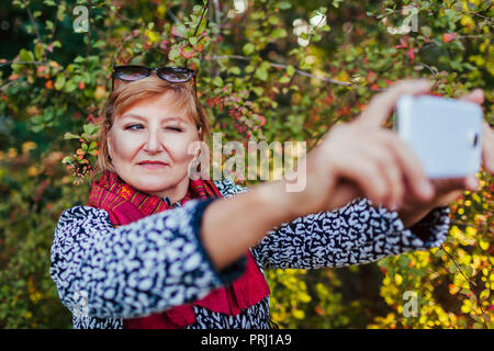 Middle-aged woman taking selfie on phone walking in park. Senior lady having fun in autumn forest Stock Photo