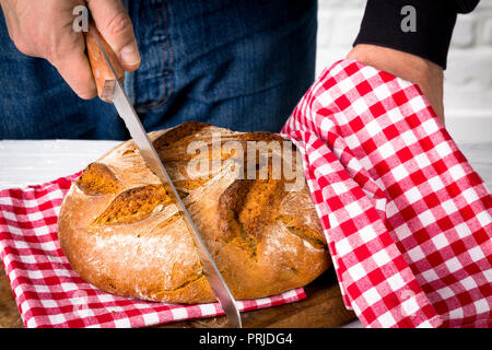 Man is cutting a loaf of bread into slices on a red and white towel an a wooden board Stock Photo