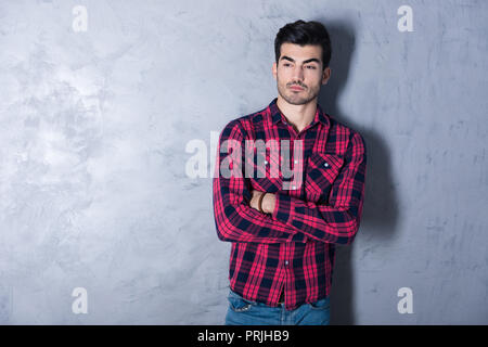 A serious handsome young man in a red checkered shirt standing in front of a grey wall in a studio. Stock Photo