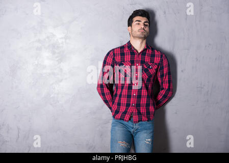 A serious handsome young man in a red checkered shirt standing in front of a grey wall in a studio. Stock Photo