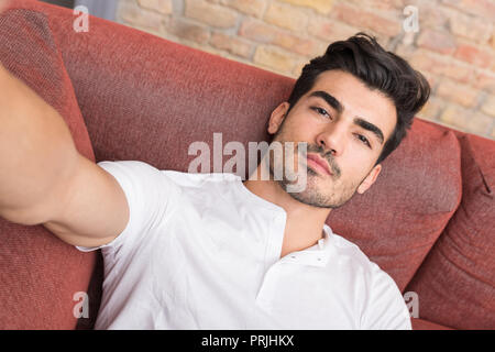 Portrait of a serious handsome young man talking a selfie while sitting on a sofa in a white tshirt from the perspective of the smartphone. Stock Photo