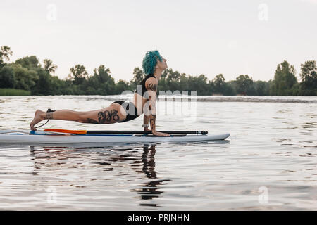 tattooed woman with blue hair practicing yoga on paddleboard in water. Upward facing dog pose (Urdhva Mukha Svanasana) Stock Photo