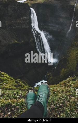 cropped shot of traveller in green boots sitting on hill in front of Haifoss waterfall Stock Photo
