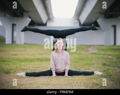 Two girls acrobats perform stand in the splits on the grass against the background of the bridge Stock Photo