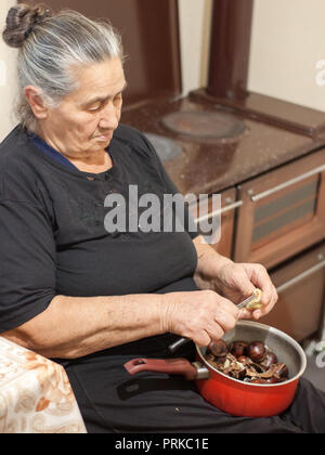 Old fashioned elder woman holding a pot with chestnuts and peeling them, next to a vintage wood heater Stock Photo