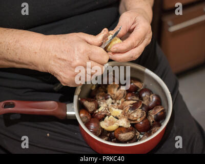 Hands of an elder woman holding a pot with chestnuts and peeling them with a knife Stock Photo