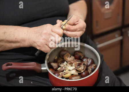 Hands of an elder woman holding a pot with chestnuts and peeling them with a knife Stock Photo