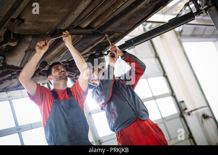Two mechanics in uniform are working in auto service with lifted vehicle Stock Photo