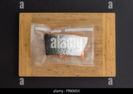 Fillet of fresh Atlantic salmon vacuum packed for freezing in clear plastic lying on a bamboo board over black in a top down view Stock Photo