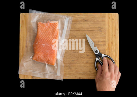Vacuum packed fillet of fresh Atlantic salmon ready for freezing or sous-vide cooking on a wooden board with a male hand reaching for a pair of scisso Stock Photo