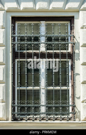 A window with a metal lattice and a wooden window frame with a cornice against a white wall. Stock Photo