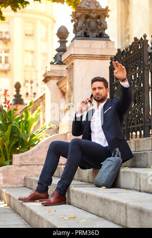A handsome young businessman sitting on the stairs talking on his smartphone and waving to someone at the Basilica in Budapest Hungary. Stock Photo