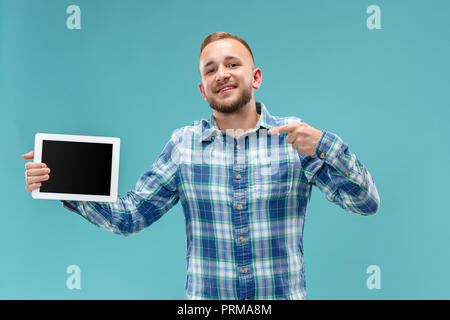 Studio picture of positive man isolated on blue background standing in casual clothes holding tablet and showing it blank screen with happy smile. He advising product or service. Stock Photo