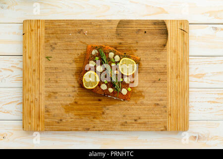 Slice of fish fillet covered in seasoning and herbs in center of wooden cutting board sitting on top of white wooden table Stock Photo