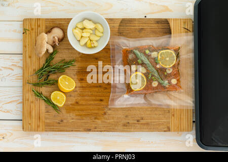 Packaged fish fillet sitting next to vacuum packer and garlic cloves inside small white bowl on top of wooden table Stock Photo