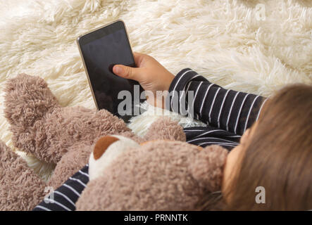 Little girl lying on the bed with her favorite teddy bear and playing a smartphone Stock Photo