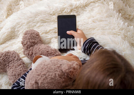 Little girl lying on the bed with her favorite teddy bear and playing a smartphone Stock Photo