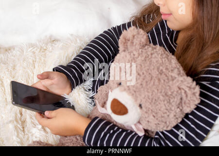 Little girl lying on the bed with her favorite teddy bear and playing a smartphone Stock Photo