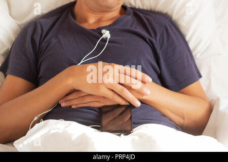 high angle view of a young caucasian man in pajamas lying down face up in bed, with his smartphone on top, with the earbuds plugged to it Stock Photo