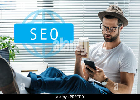 bearded developer in hat holding coffee to go and using smartphone with SEO sign Stock Photo
