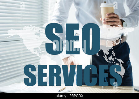 cropped view of developer holding laptop and coffee to go, with SEO services and world map Stock Photo