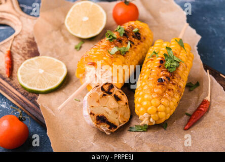 Grilled corn cobs. Delicious summer snack. With parsley, chili peppers, lime and grilled garlic. Served on old wooden cutting board with parchment pap Stock Photo
