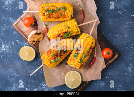 Grilled corn cobs. Delicious summer snack. With parsley, chili peppers, lime and grilled garlic. Served on old wooden cuttingg board with parchment pa Stock Photo