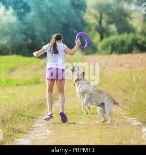 A young girl with a dog runs along a country road in a field in summer. The girl holds in her hand a ring for training Stock Photo