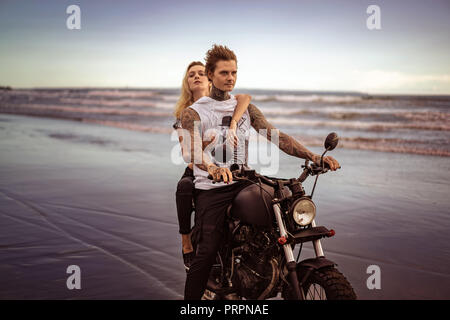 young stylish tattooed couple riding motorcycle on ocean beach Stock Photo