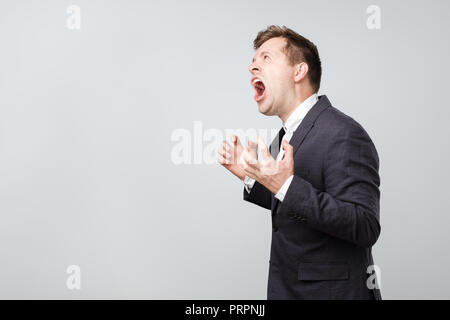 Side view of young caucasian man screaming loudly while standing isolated on gray background with copyspace. Stock Photo