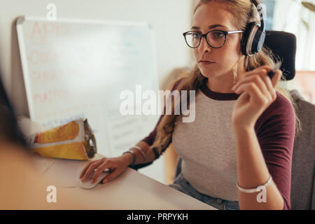 Young caucasian woman wearing headphones sitting at her desk and working on computer. Female programmer working on new software development. Stock Photo