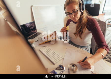 Woman writing programme codes on paper and eating. Female coder working on new software development at tech startup in office. Stock Photo