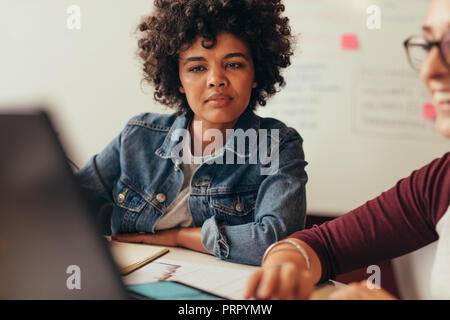 African woman looking at her colleagues laptop screen while working. Young people working at small coworking space in office. Stock Photo