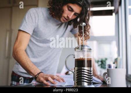 Young man reading book standing in kitchen at home while making coffee. Student standing in kitchen and studying for college exam. Stock Photo