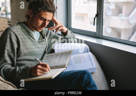 Student doing his home work sitting on couch beside a window. Man writing in book sitting at home with focus and concentration. Stock Photo