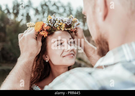 Loving bearded man putting beautiful floral chaplet on head of his appealing woman Stock Photo