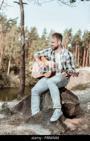 Handsome bearded man wearing jeans and squared shirt playing the guitar near lake Stock Photo